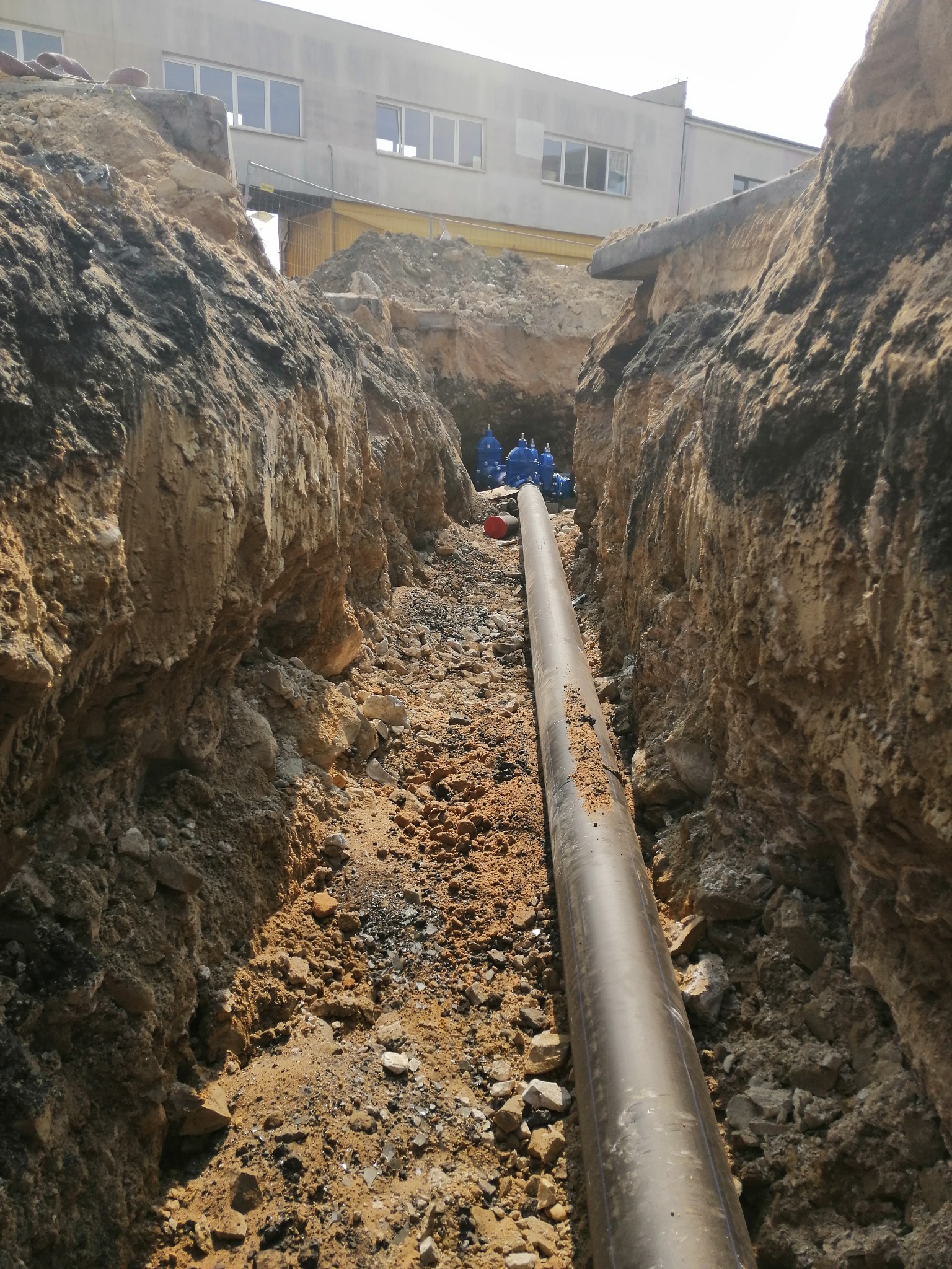 Rura ułożona w wykopie ziemnym, widoczni pracownicy w oddali, budynek w tle.