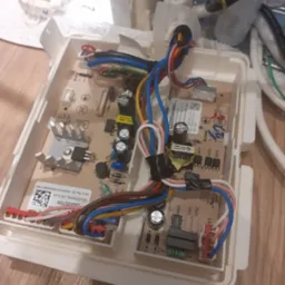 Otwarta obudowa modułu sterującego urządzenia AGD z widoczną płytką drukowaną, przewodami w różnych kolorach i elementami elektronicznymi. Widoczne ślady naprawy w postaci taśmy izolacyjnej.