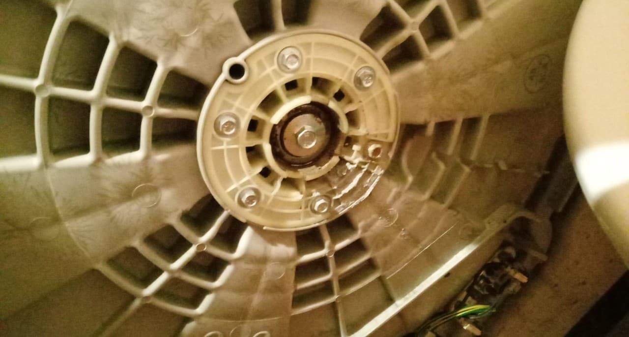Wnętrze pralki automatycznej z widocznym uszkodzonym mocowaniem bębna, centralnie umieszczony element mocujący z sześcioma śrubami i śladami korozji.