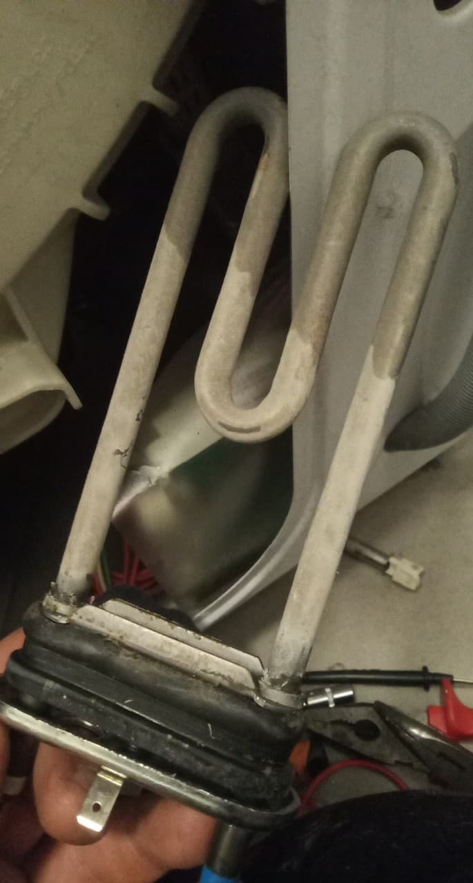 Wyjęta, zakamieniona grzałka z uszczelką i elementami mocującymi, widoczna w dłoni, obok narzędzi, prawdopodobnie z pralki.