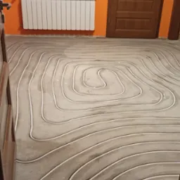 Białe rurki ułożone spiralnie w szarej wylewce podłogowej, widoczny grzejnik na pomarańczowej ścianie i fragmenty drewnianych drzwi.