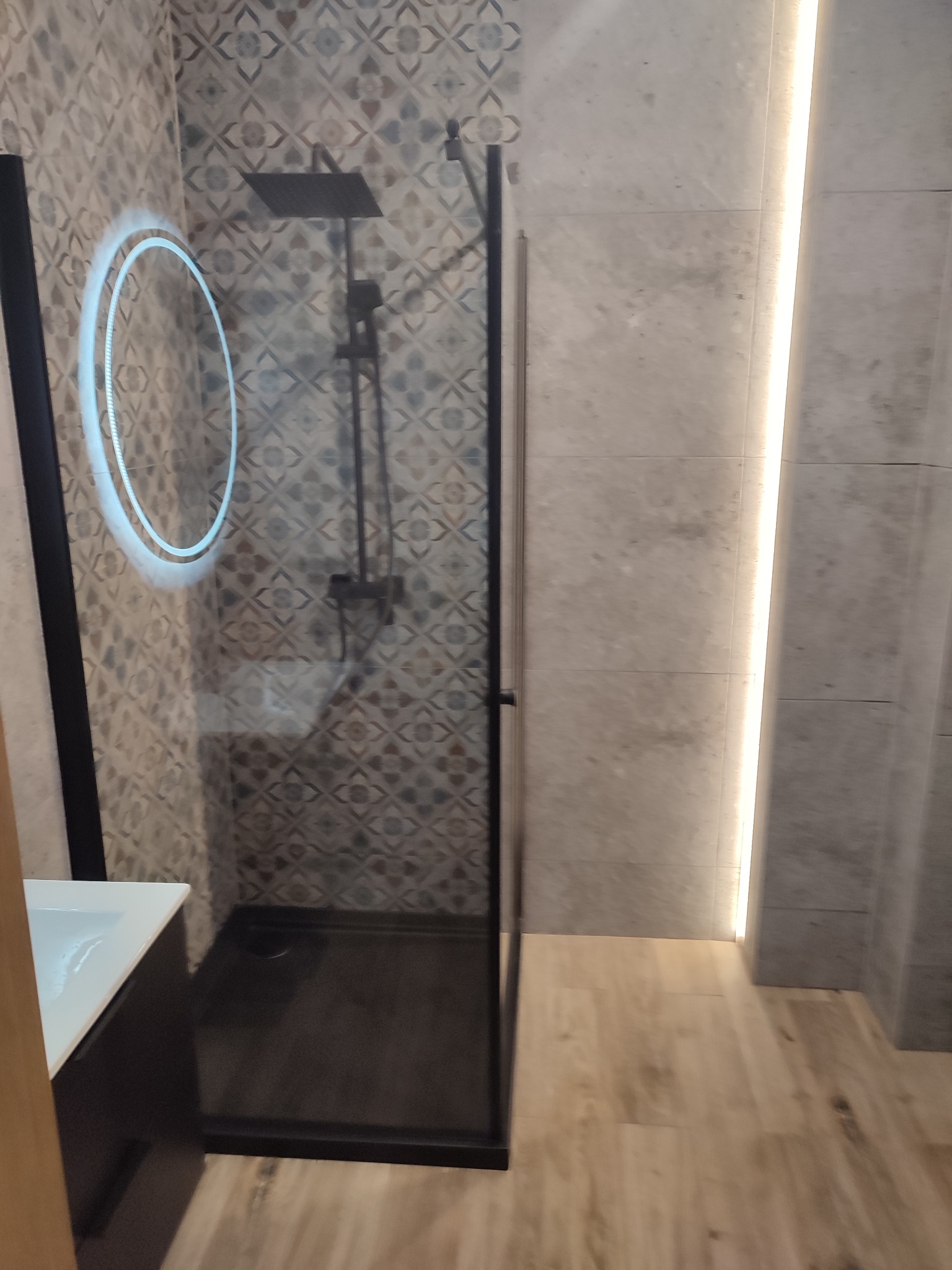 Nowoczesna łazienka z kabiną prysznicową typu walk-in z czarnymi profilami, okrągłym lustrem LED i płytkami w geometryczne wzory na ścianie.