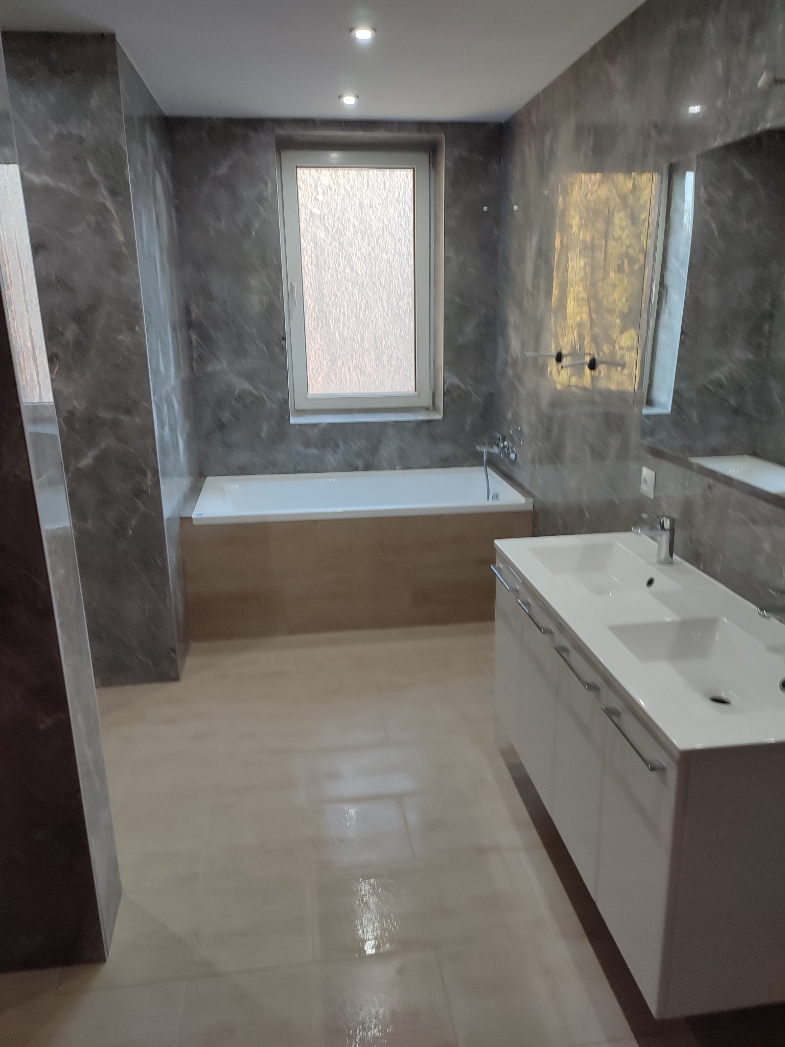 Nowoczesna łazienka z marmurowymi ścianami, białą wanną z drewnianą obudową i podwójną umywalką z szafką, oświetlona punktowymi lampami sufitowymi.