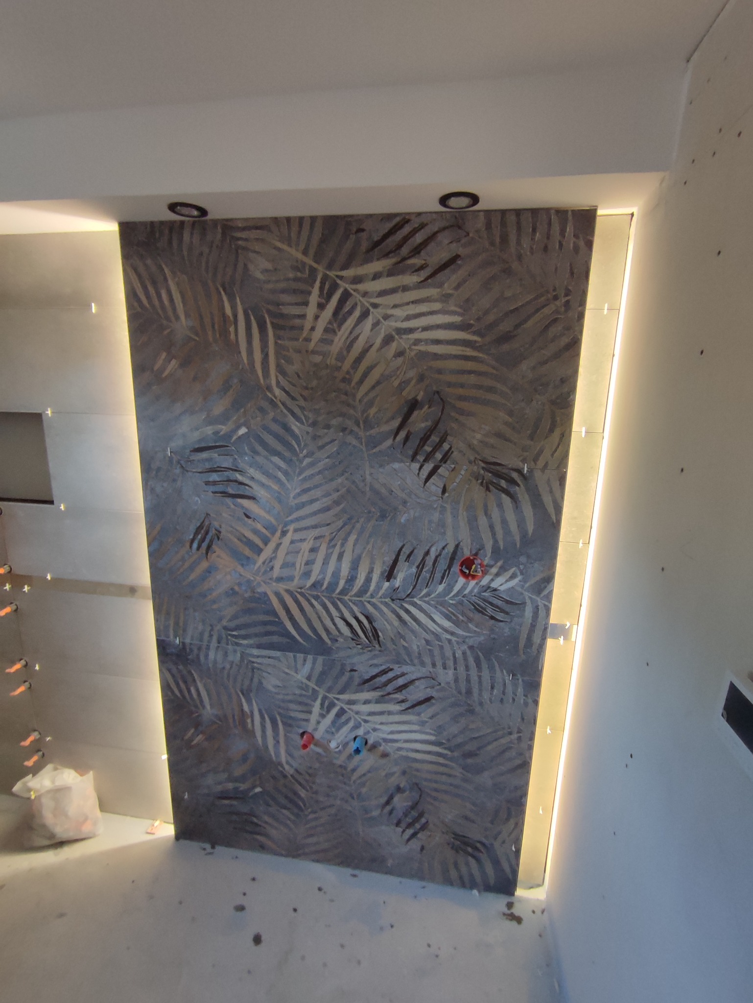 Fragment ściany z ułożonymi płytkami z motywem liści, podświetlony z boku, widoczne wystające rurki instalacji wodnej, w tle worki z materiałami budowlanymi.