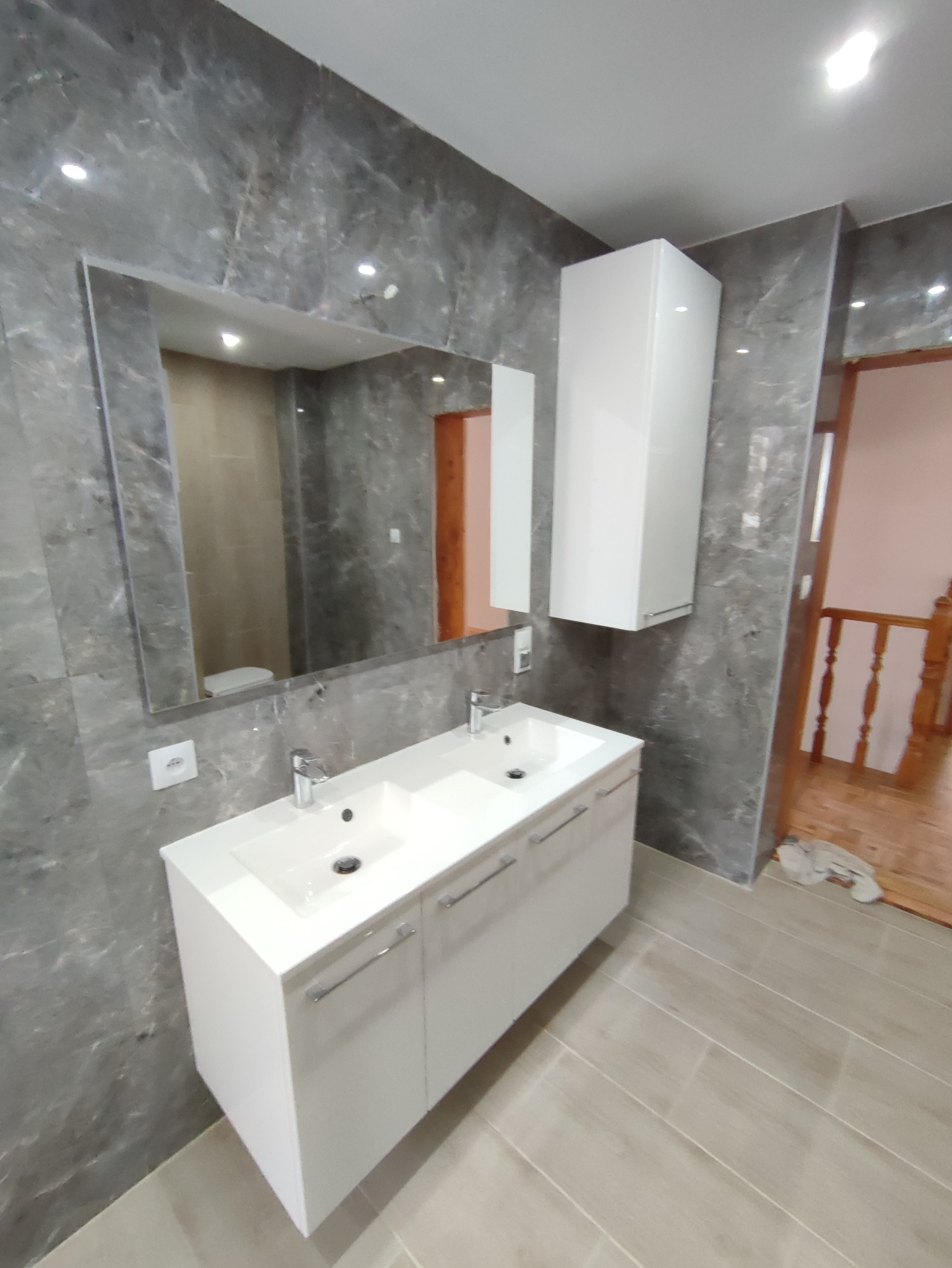 Nowoczesna łazienka z umywalką nablatową z dwiema misami, dużym lustrem i szarą marmurową okładziną ścian. Widoczna szafka wisząca i fragment drewnianych schodów w tle.