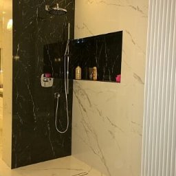 Dawid Warzała Usługi Remontowe - Nowoczesna łazienka z czarną marmurową ścianą prysznicową, wnęką na kosmetyki i białymi płytkami imitującymi marmur na pozostałych ścianach i podłodze.