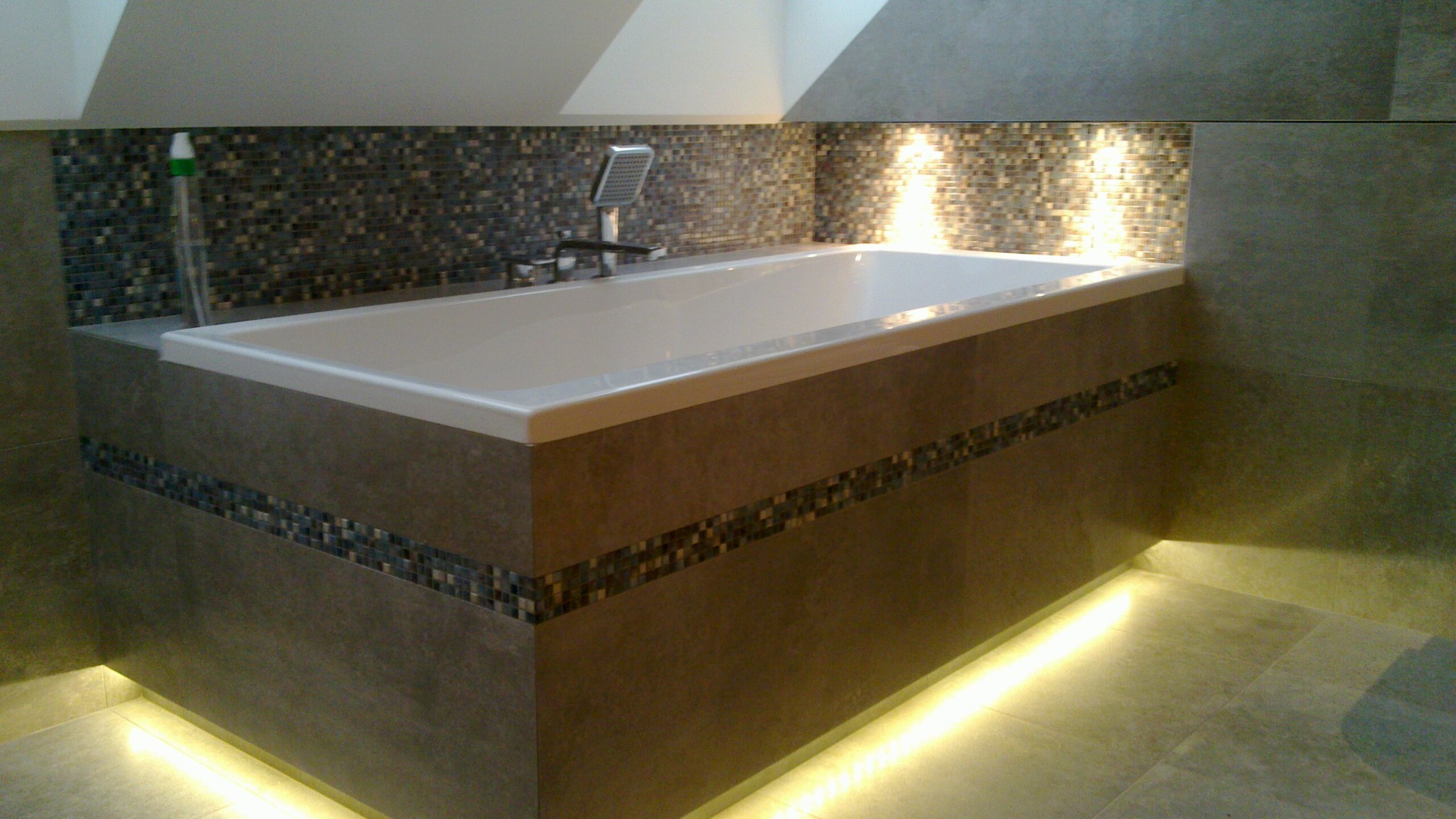 Nowoczesna wanna obudowana płytkami w odcieniach szarości, z dekoracyjnym pasem mozaiki i subtelnym podświetleniem LED u dołu, uzupełnona armaturą i mozaiką na ścianie.