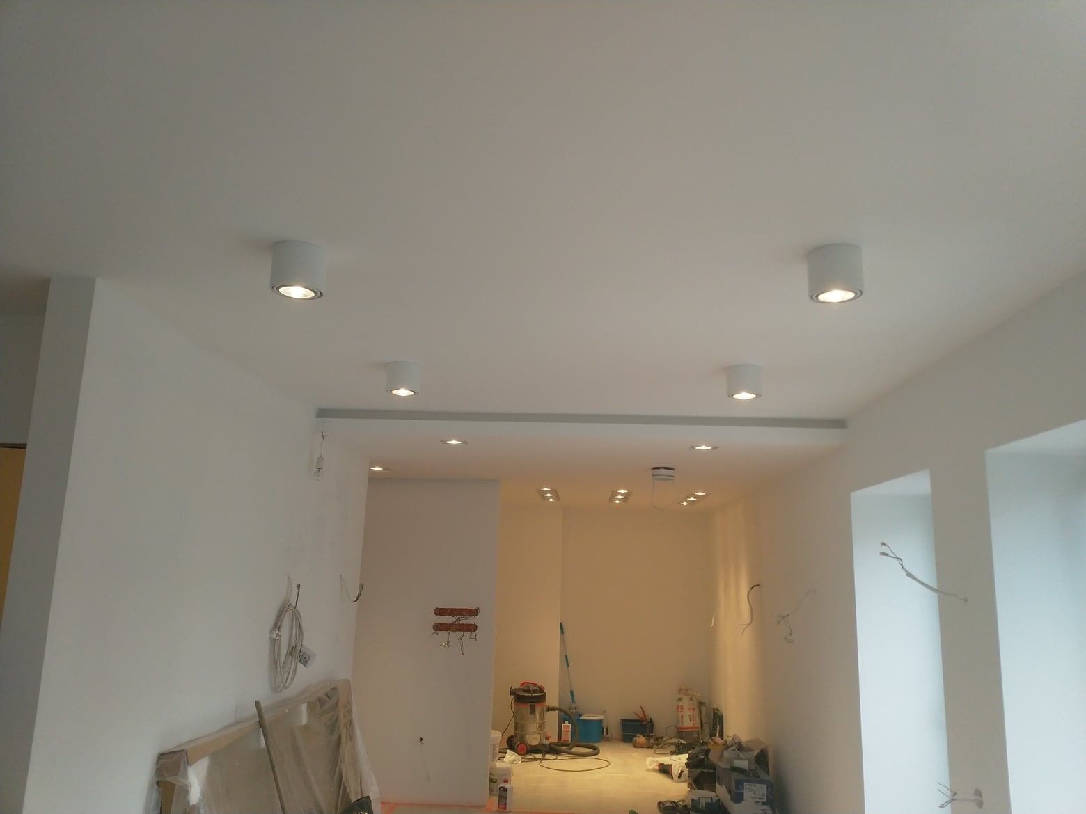 Wnętrze w trakcie prac wykończeniowych, widoczne białe ściany, sufit z zamontowanym oświetleniem punktowym, przygotowane materiały i narzędzia budowlane na podłodze.