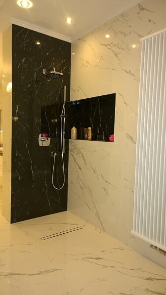 Nowoczesna łazienka z czarną marmurową ścianą prysznicową, wnęką na kosmetyki i białymi płytkami imitującymi marmur na pozostałych ścianach i podłodze.