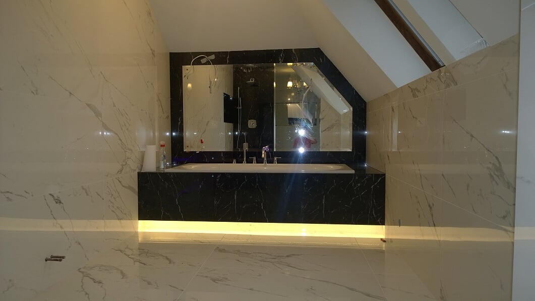 Nowoczesna łazienka na poddaszu z wanną obudowaną czarnym marmurem, duże lustro w czarnej ramie, białe płytki imitujące marmur na ścianach i podłodze, oświetlenie LED pod wanną.