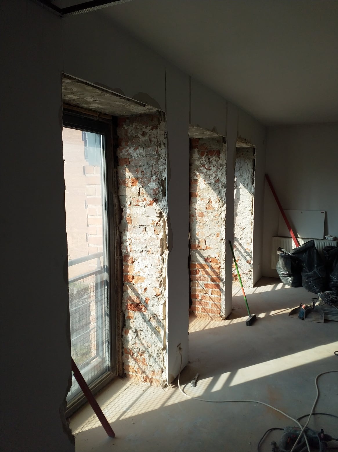 Wnętrze w trakcie prac przygotowawczych, odsłonięta cegła na filarach, widoczne ślady po kuciu, częściowo zagruntowane ściany, podłoga przygotowana do dalszych prac.