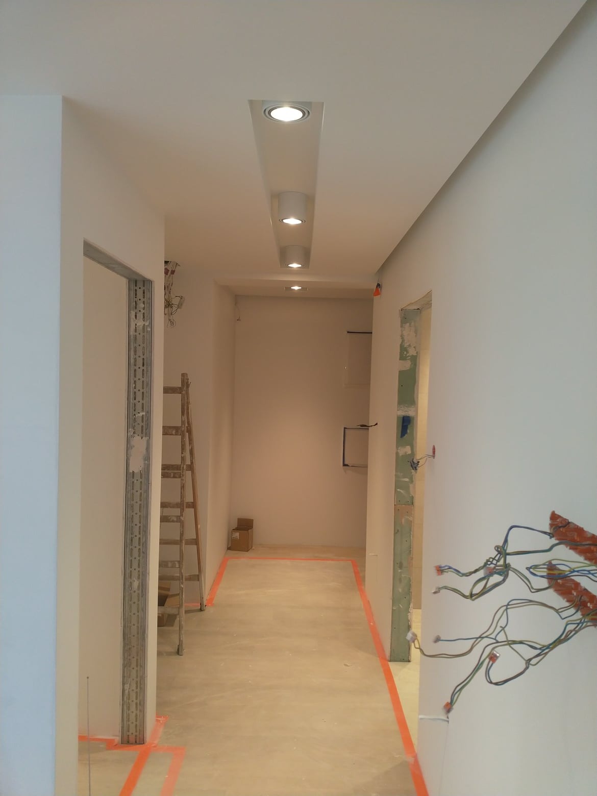 Korytarz w trakcie prac wykończeniowych, widoczne przewody elektryczne, drabina i oświetlenie sufitowe w zabudowie.