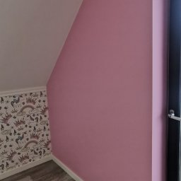 GOLD-MAL Marcin Białobłocki - Pomalowane na różowo poddasze z oknem, drewnopodobną podłogą i fragmentem ściany z tapetą w jednorożce i korony.