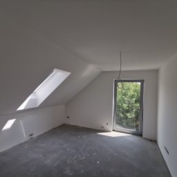 GOLD-MAL Marcin Białobłocki - Poddasze po szpachlowaniu i malowaniu na biało, widoczne okno dachowe i duże okno z widokiem na zieleń, surowa podłoga przed wykończeniem.