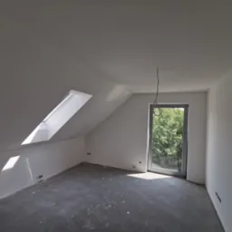 Poddasze po szpachlowaniu i malowaniu na biało, widoczne okno dachowe i duże okno z widokiem na zieleń, surowa podłoga przed wykończeniem.