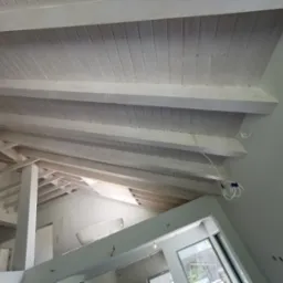Poddasze z drewnianą konstrukcją stropu, pomalowaną na biało, widok z dołu w trakcie prac wykończeniowych, widoczne kable oświetleniowe, ściany w jasnym odcieniu szarości.
