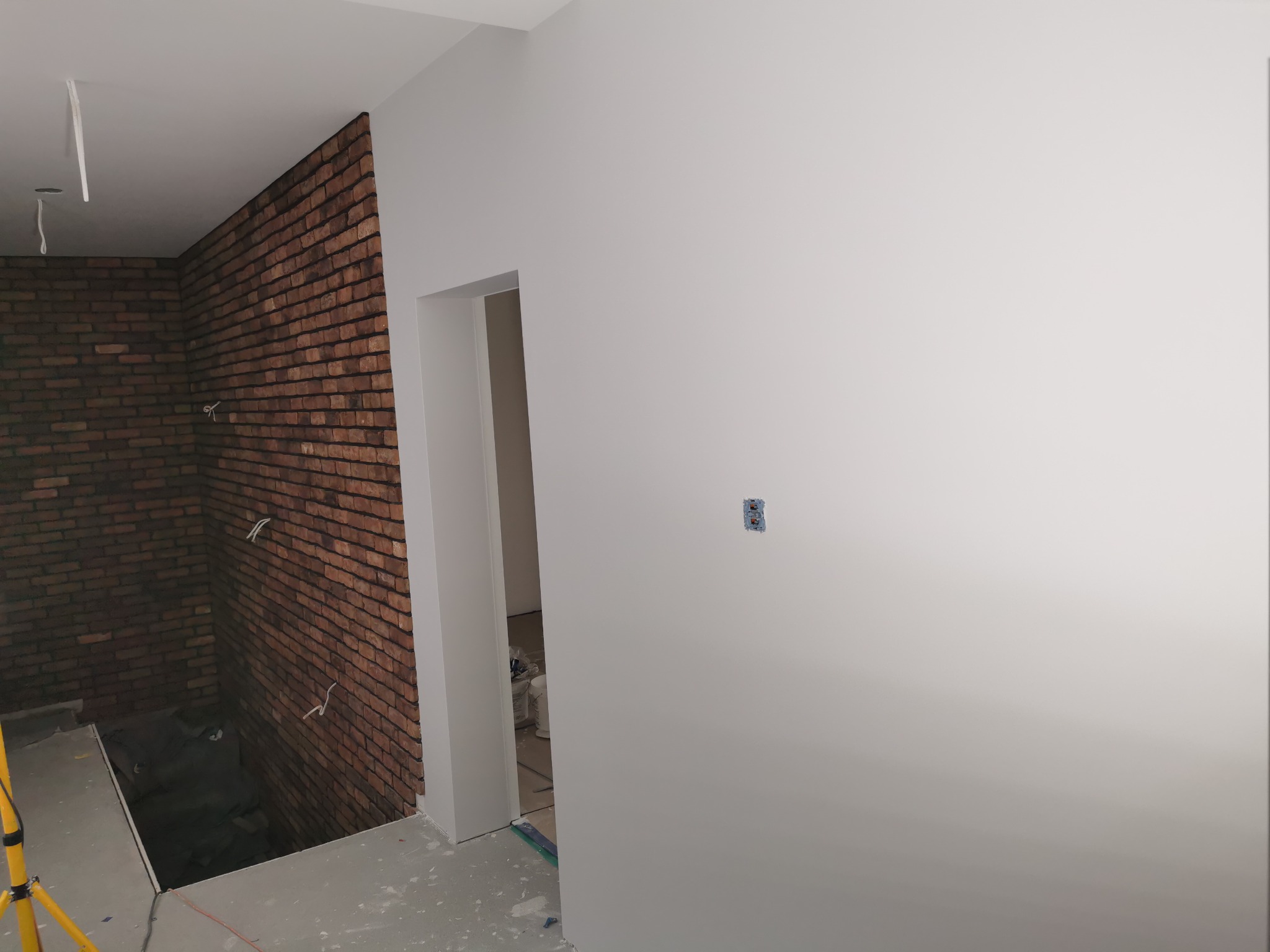 Wnętrze w trakcie prac wykończeniowych: pomalowana na biało ściana z widocznym gniazdkiem, fragment ściany wyłożonej cegłą klinkierową, otwarte przejście do innego pomieszczenia, widoczne kable...