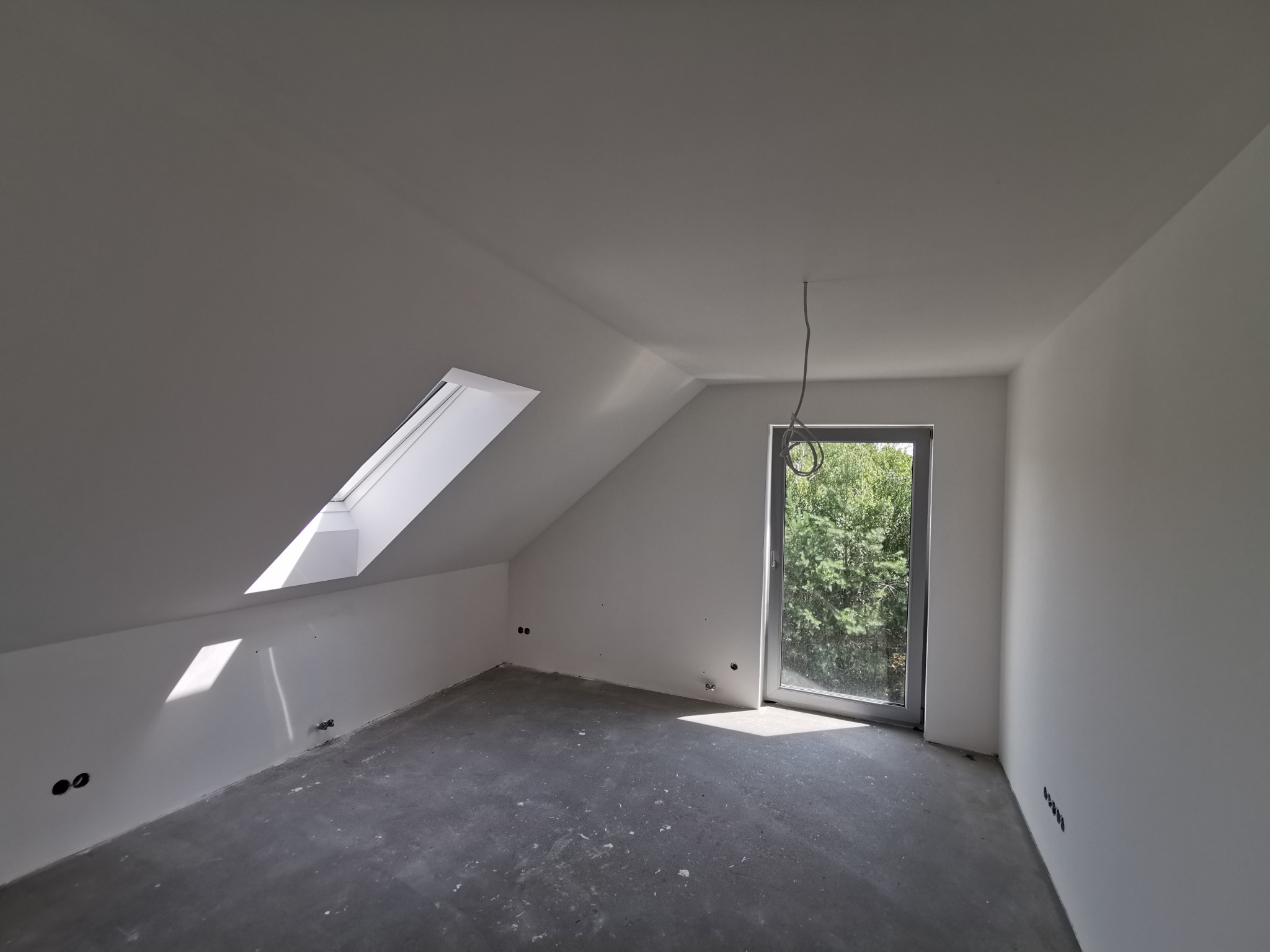 Poddasze po szpachlowaniu i malowaniu na biało, widoczne okno dachowe i duże okno z widokiem na zieleń, surowa podłoga przed wykończeniem.