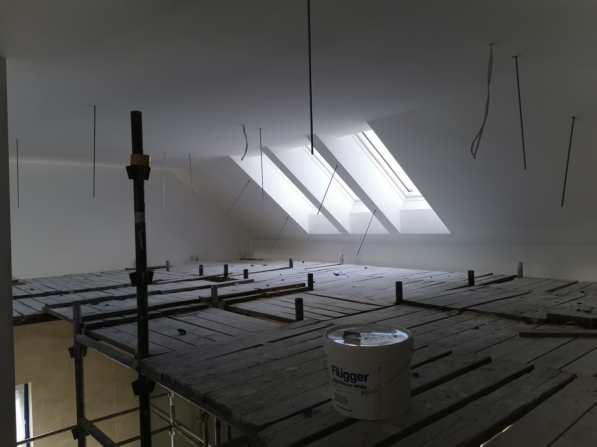 Poddasze przygotowywane do prac wykończeniowych z widocznym rusztowaniem, wiadrem z gruntem Flugger Wall Primer White oraz wystającymi przewodami z sufitu w pobliżu okien dachowych.