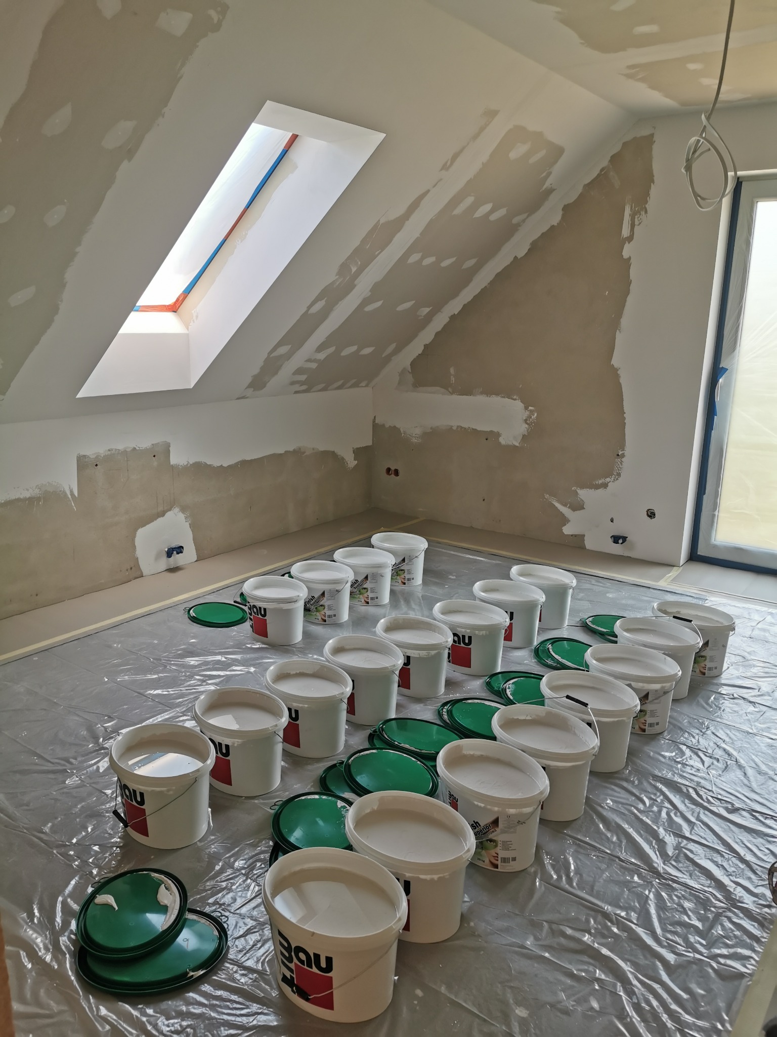 Poddasze w trakcie prac przygotowawczych do malowania: ściany częściowo pokryte gładzią, podłoga zabezpieczona folią, widoczne liczne wiadra z farbą i otwarte wieczka.