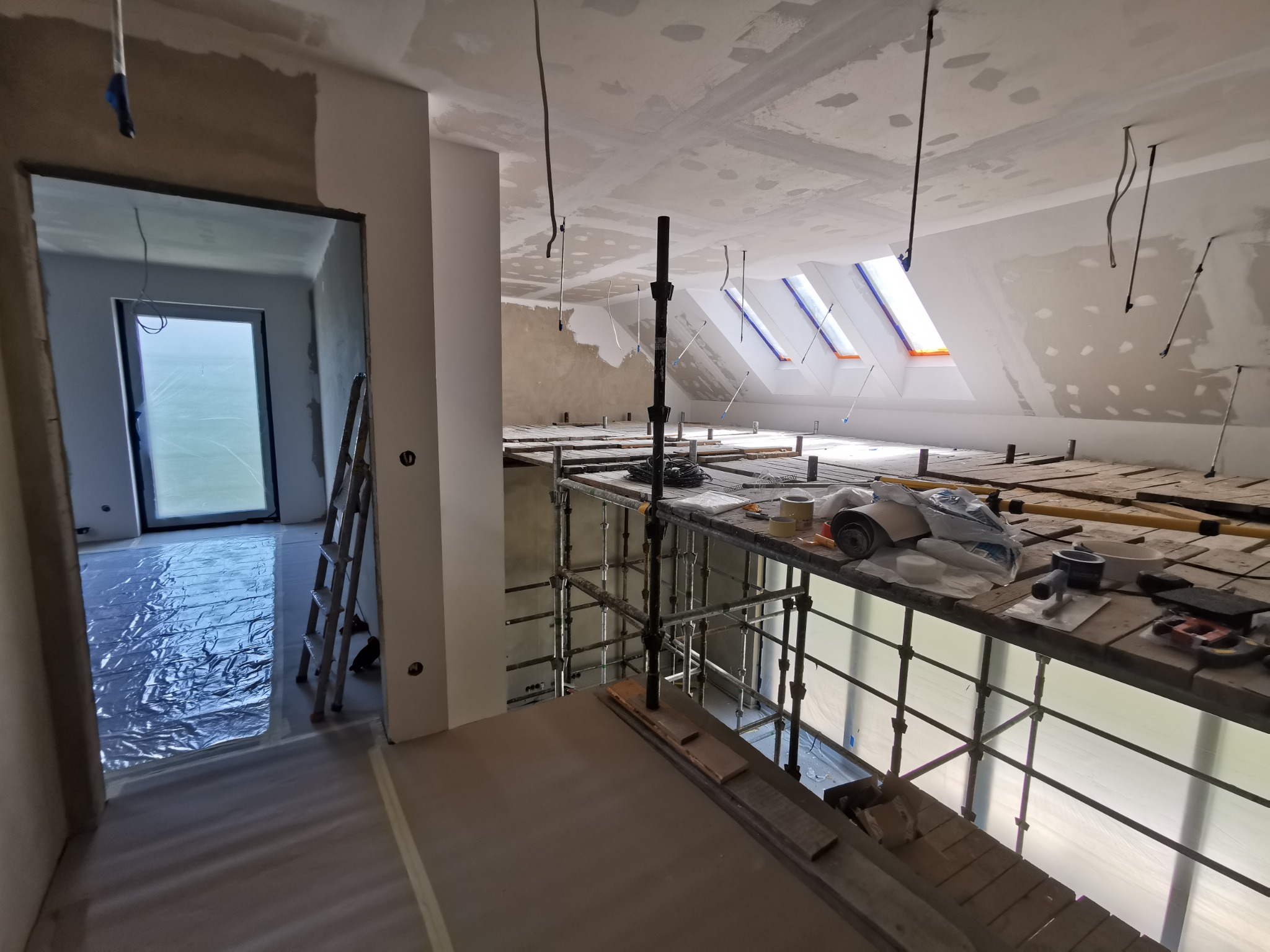 Poddasze w trakcie prac wykończeniowych: widoczne rusztowanie, okna dachowe z folią ochronną, ściany przygotowane pod malowanie i instalacje elektryczne wystające z sufitu.