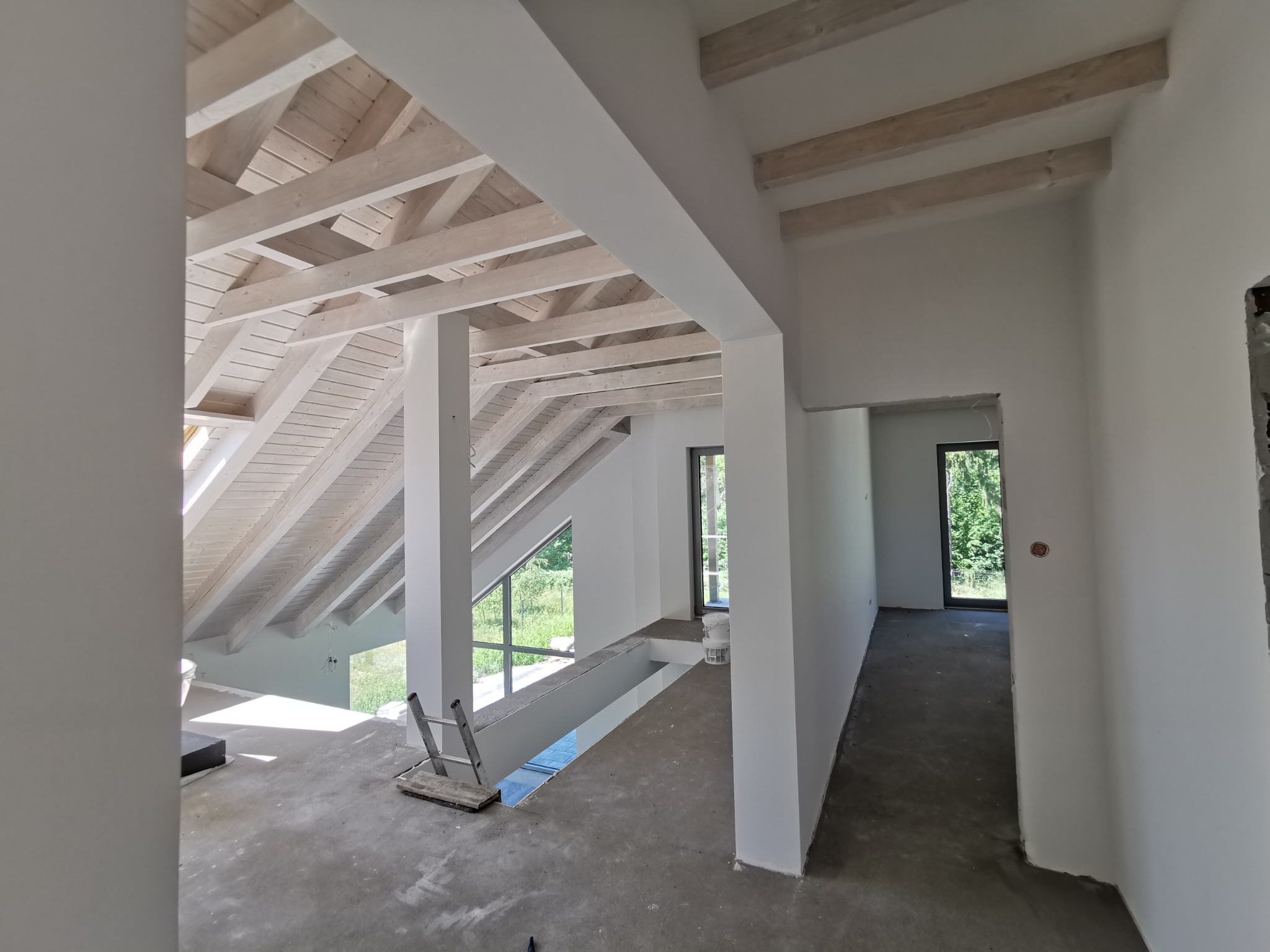 Wnętrze domu w trakcie prac wykończeniowych: pomalowane ściany na biało, drewniana konstrukcja dachu również w bieli, surowa betonowa podłoga, okna z widokiem na zieleń, drabina oparta o ścianę...
