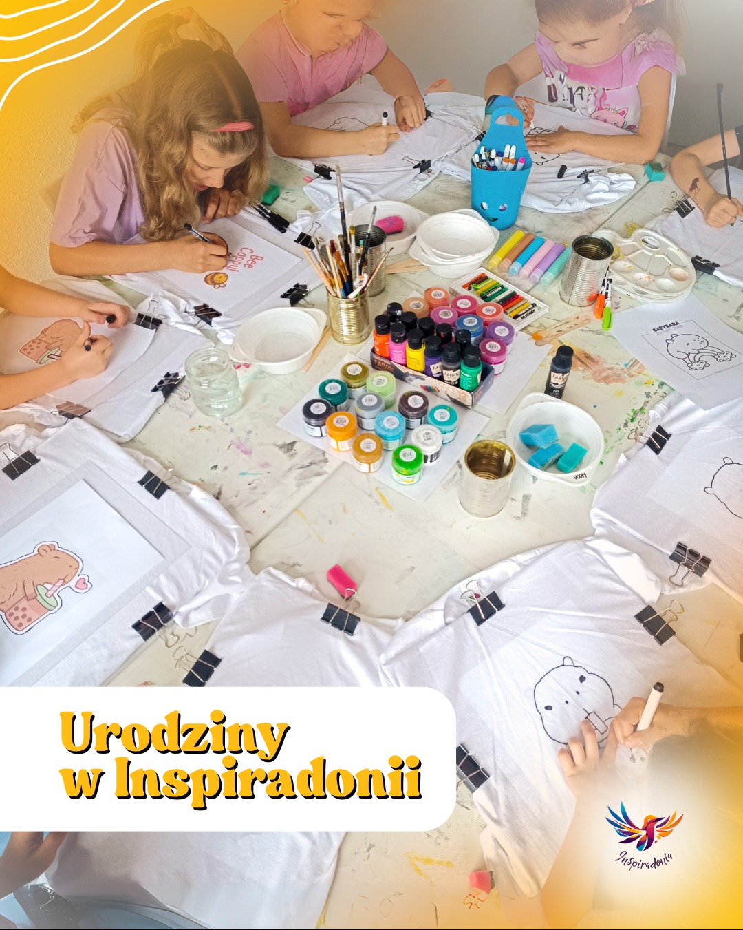 Dzieci dekorują białe koszulki farbami na stole podczas warsztatów artystycznych. Widoczne pędzle, farby, szablony i logo 'Inspiradonii'.
