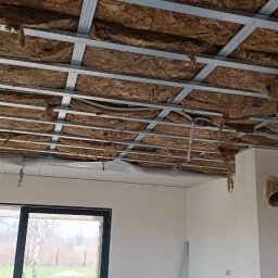 Paweł Radzymiński - Montaż sufitu podwieszanego z izolacją z wełny mineralnej, widoczne profile metalowe i instalacja elektryczna w rurkach karbowanych, w tle okno i materiały budowlane.