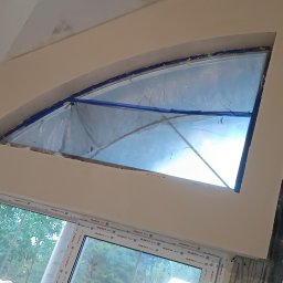 Paweł Radzymiński - Przygotowanie do prac wykończeniowych: okno zabezpieczone folią i taśmą malarską, ściana po nałożeniu gładzi, surowy mur z betonu komórkowego w tle.