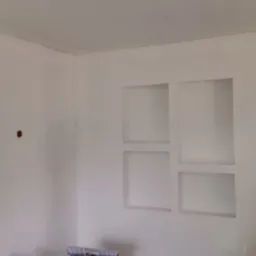 Świeżo pomalowane na biało wnętrze z wnęką na ścianie, widoczne puszki elektryczne i rolka tapety na podłodze.