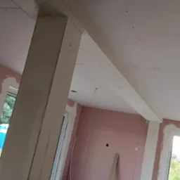 Wnętrze w trakcie prac wykończeniowych: ściany obłożone płytami karton-gips, widoczna konstrukcja sufitu podwieszanego, narzędzia i materiały budowlane na podłodze.