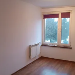 Pusty pokój z białymi ścianami, brązowymi panelami podłogowymi, grzejnikiem i oknem z bordowymi roletami.