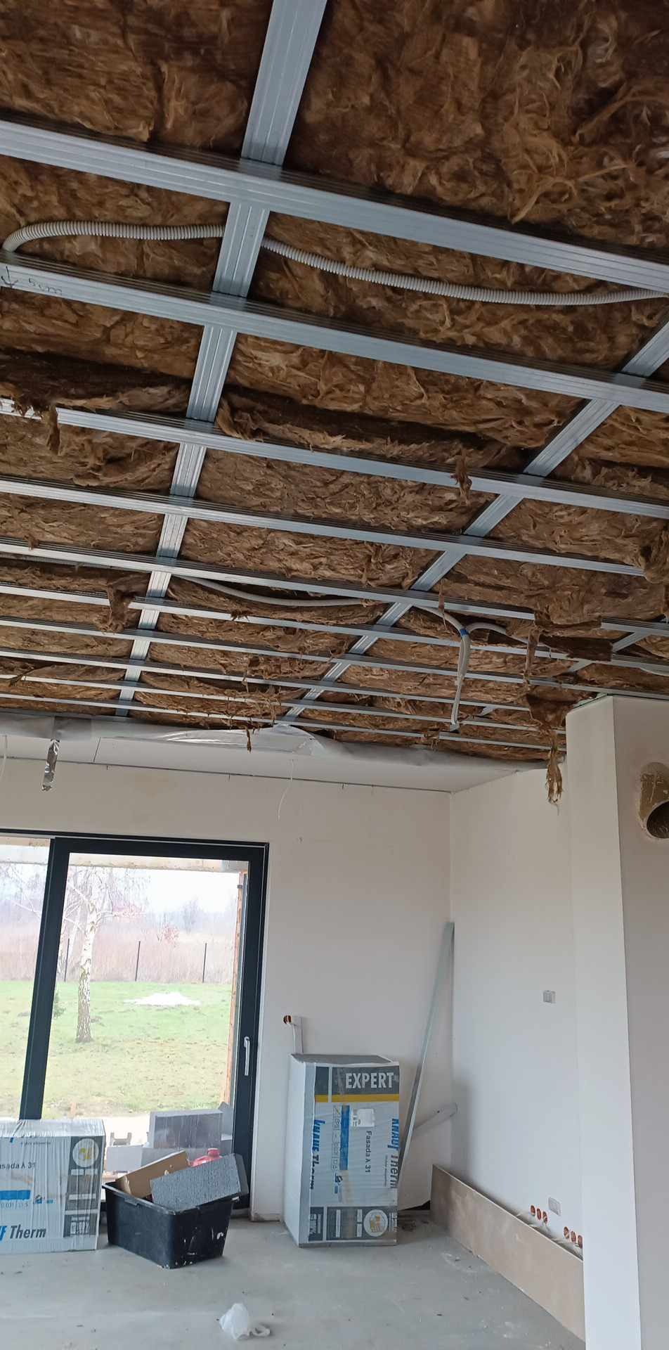 Montaż sufitu podwieszanego z izolacją z wełny mineralnej, widoczne profile metalowe i instalacja elektryczna w rurkach karbowanych, w tle okno i materiały budowlane.
