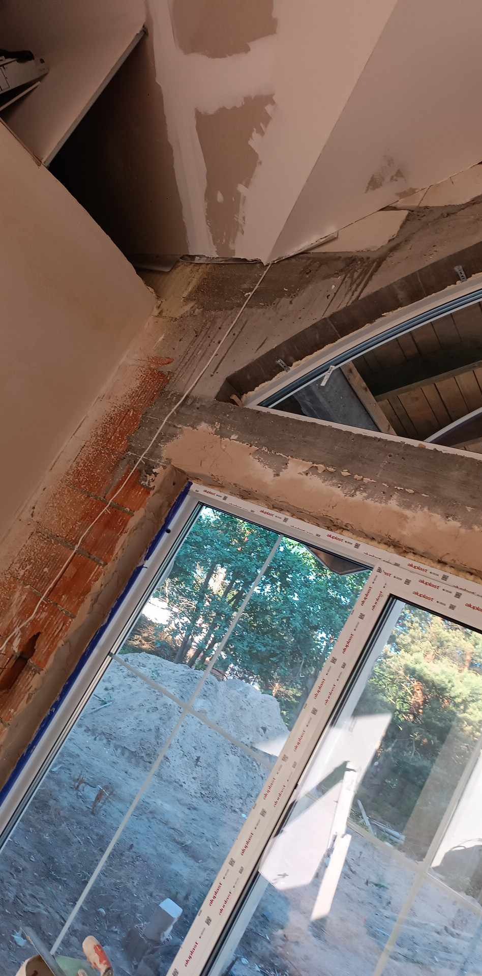 Wnętrze w trakcie prac wykończeniowych, widoczne surowe ściany z fragmentami cegieł, niedokończone gładzie na suficie i ścianach, nowe okno PCV z widokiem na zewnątrz.