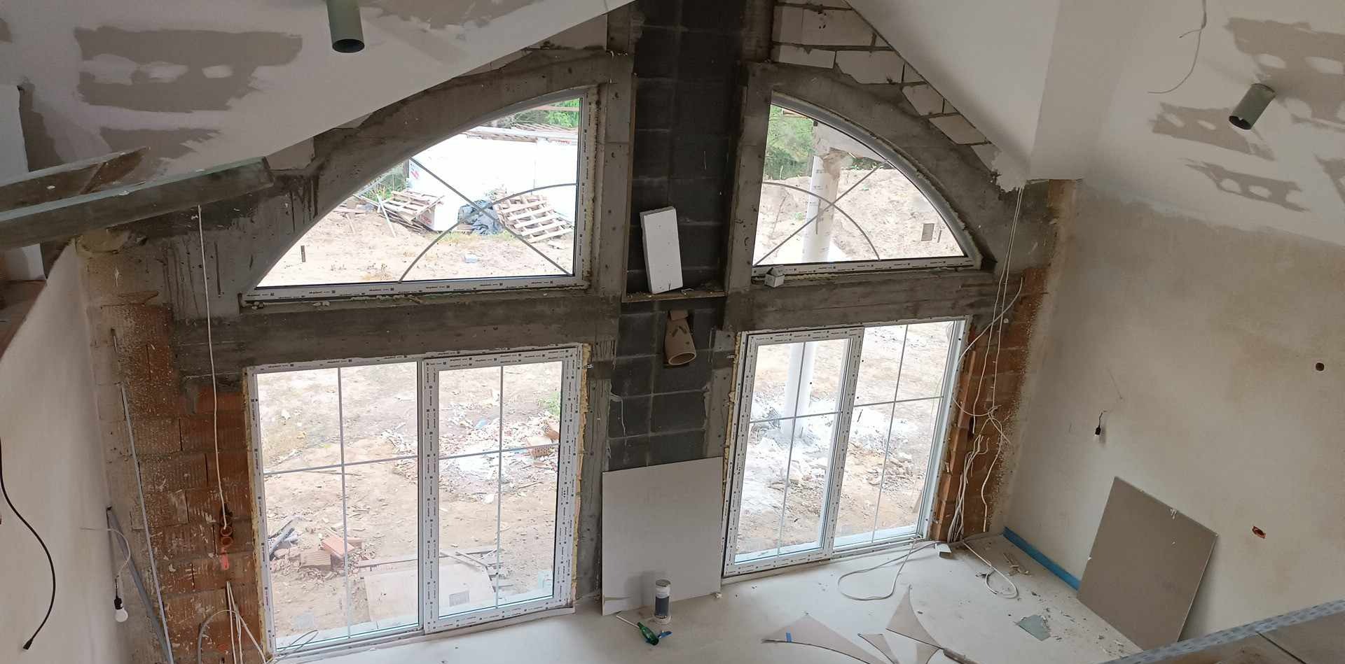 Wysoki pokój na poddaszu w stanie surowym, z dużymi oknami o nietypowym kształcie, widoczne ślady po szpachlowaniu na ścianach i suficie, wystające kable elektryczne.