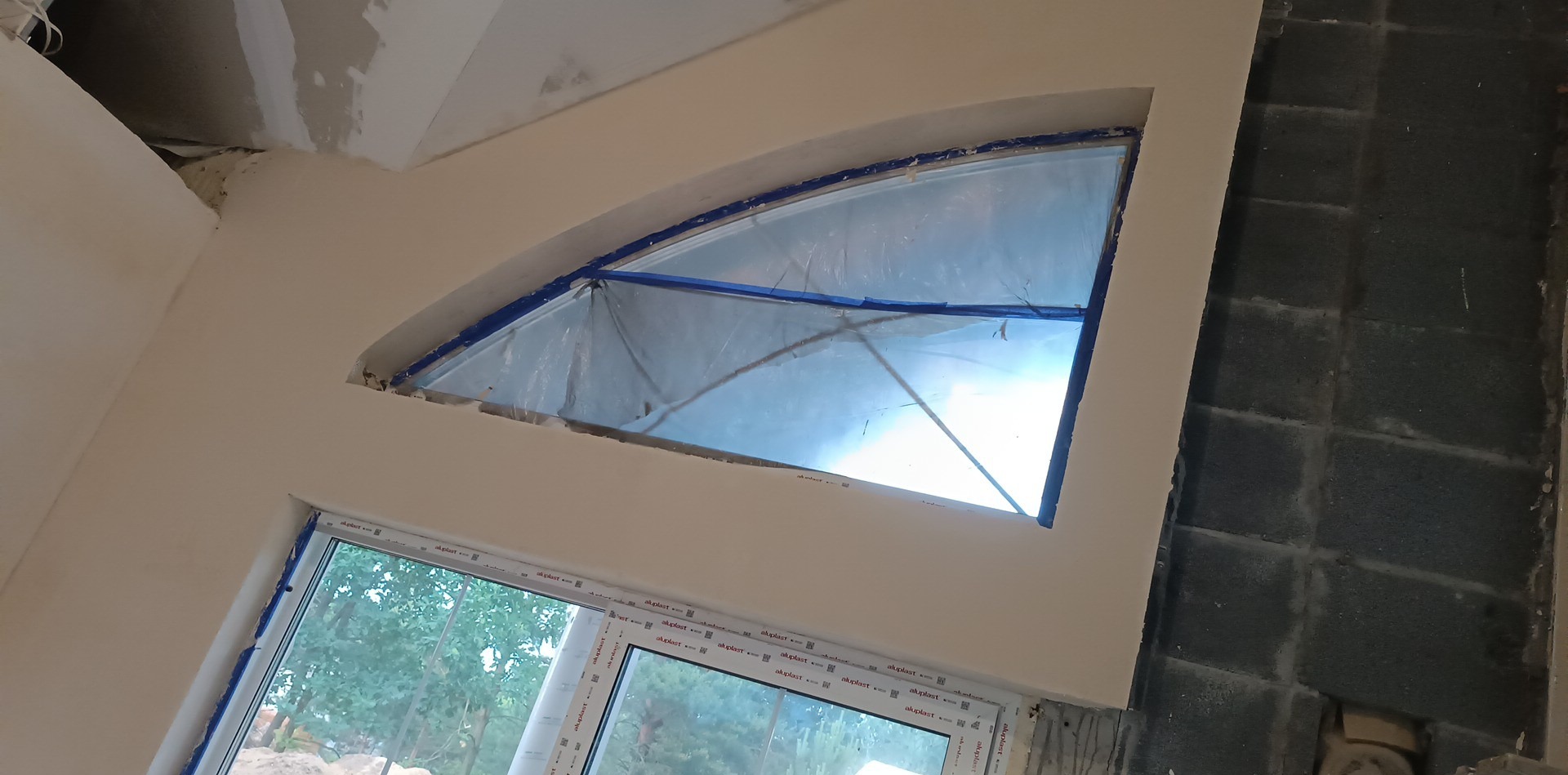 Przygotowanie do prac wykończeniowych: okno zabezpieczone folią i taśmą malarską, ściana po nałożeniu gładzi, surowy mur z betonu komórkowego w tle.