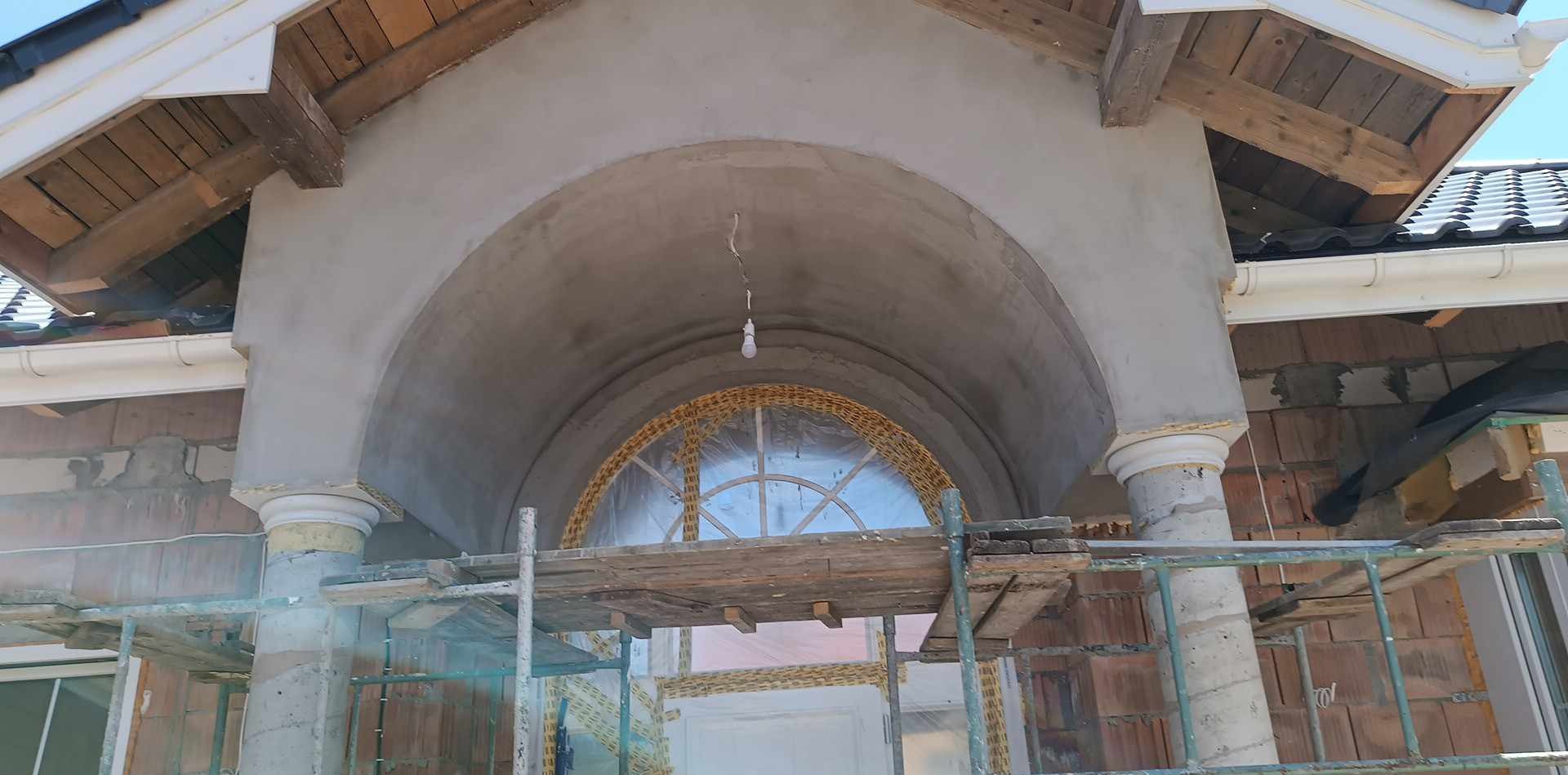 Łukowate wejście do budynku w trakcie wykonywania, widoczne rusztowanie, cegły i częściowo nałożona warstwa szarej zaprawy.