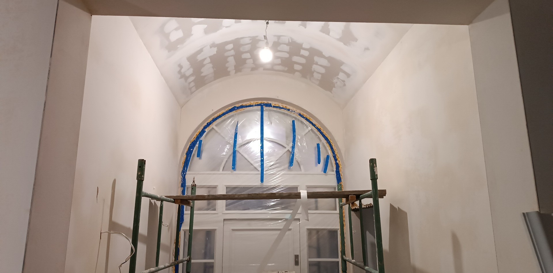 Korytarz w trakcie prac wykończeniowych: ściany pokryte gładzią, sufit łukowy z widocznymi łatami, oklejone folią drzwi łukowe, rusztowanie w dolnej części kadru.