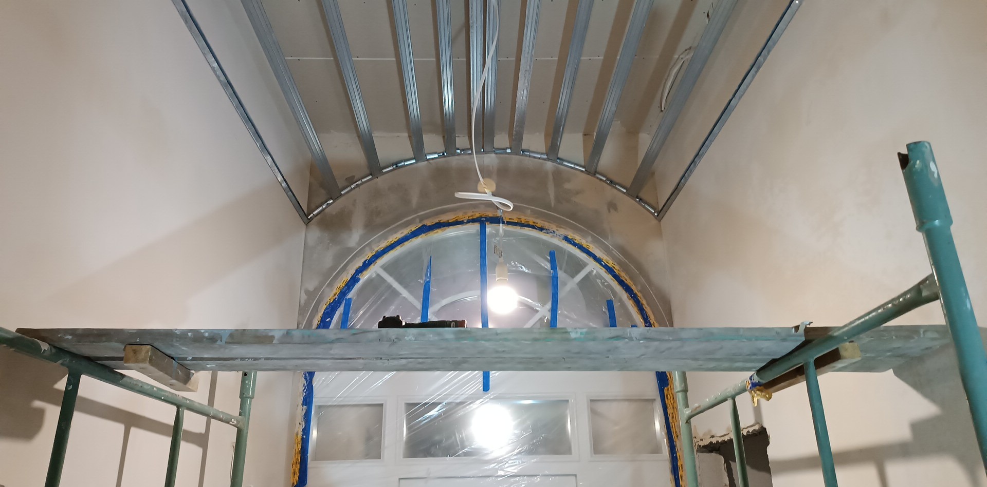 Montaż metalowej konstrukcji sufitu podwieszanego z płyt kartonowo-gipsowych, widoczne rusztowanie i okno zabezpieczone folią, w tle ściana przygotowana do dalszych prac wykończeniowych.