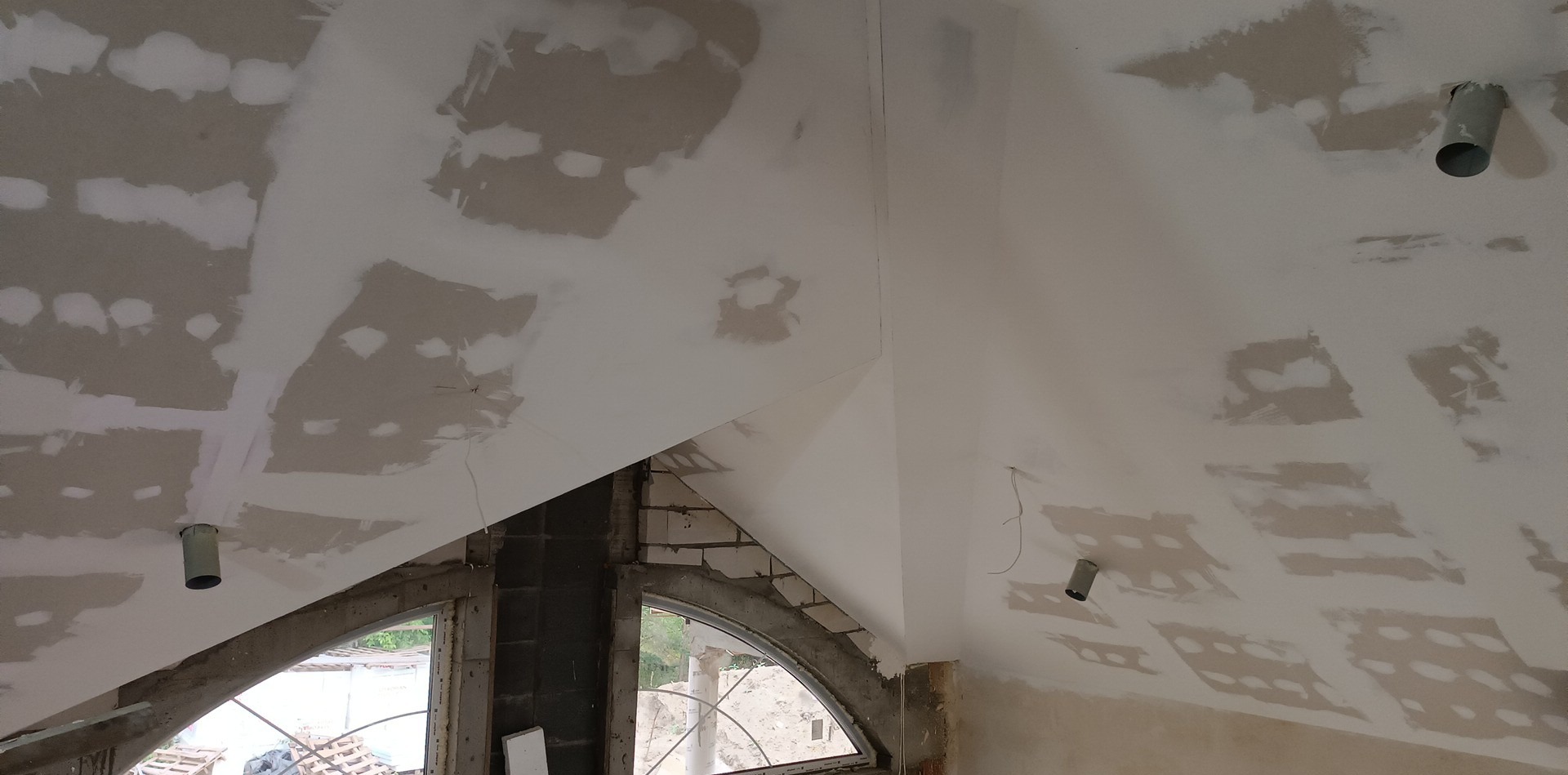 Sufit poddasza z oknem łukowym, częściowo pokryty gładzią, widoczne niedoskonałości i wystające rury instalacyjne.