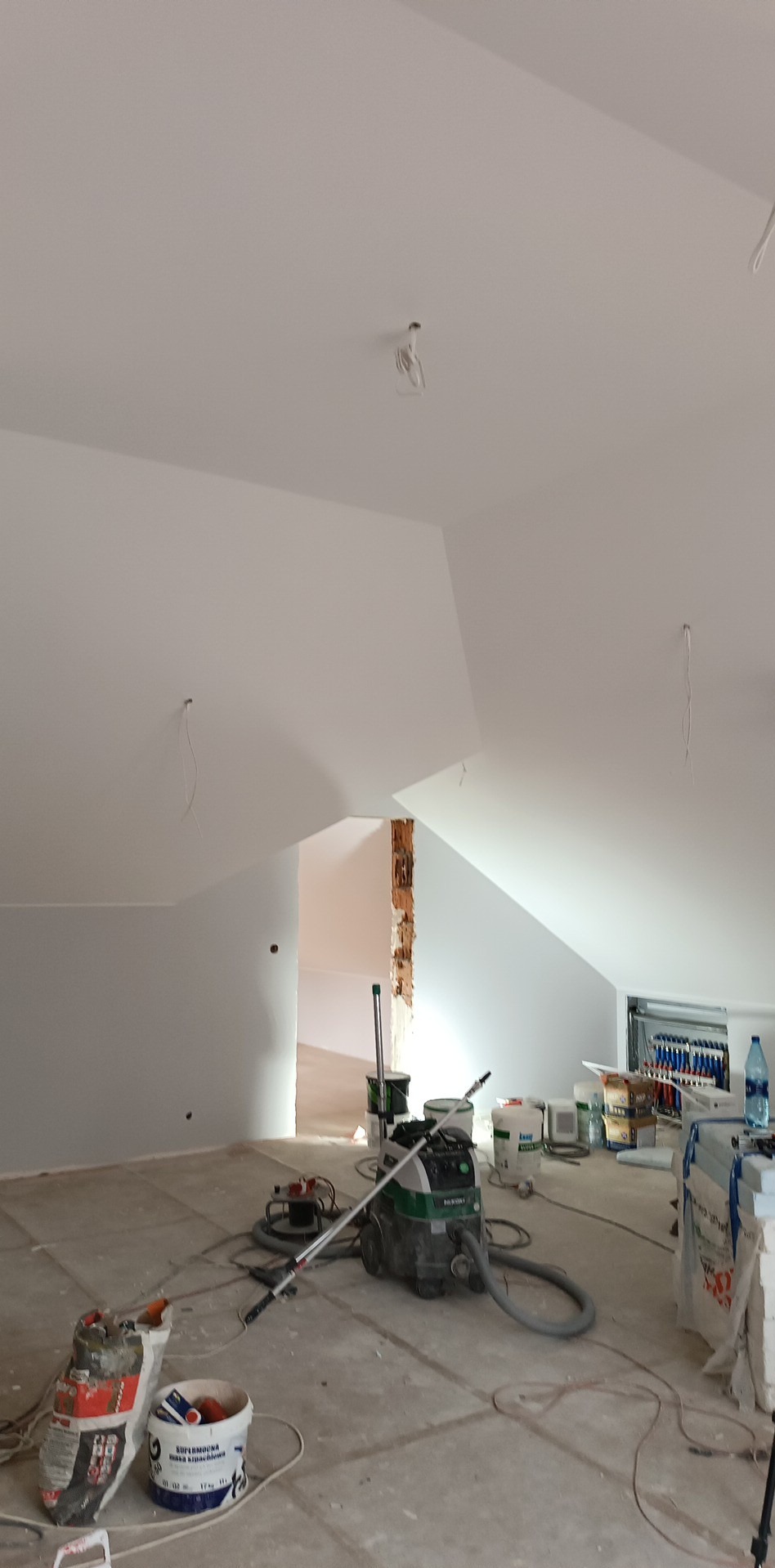 Pokój na poddaszu w trakcie prac wykończeniowych, widoczny agregat szpachlarski, worki z zaprawą i wiaderka z masą szpachlową na podłodze, ściany i sufit pomalowane na biało, widoczne przewody...
