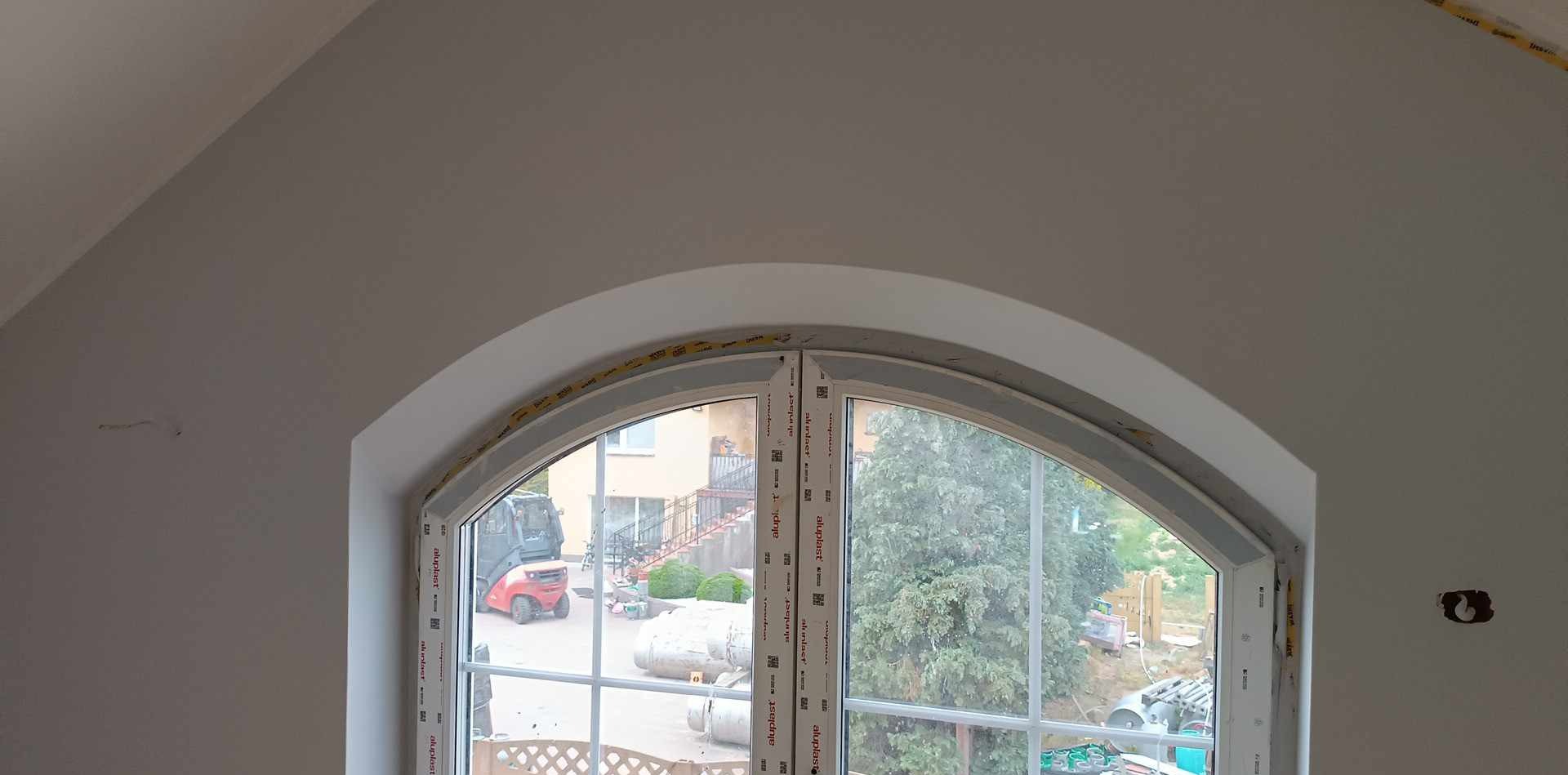 Wnętrze z zaokrąglonym oknem, ściany przygotowane do dalszych prac wykończeniowych, widoczne ślady po instalacji elektrycznej, za oknem widok na podwórko z wózkiem widłowym.