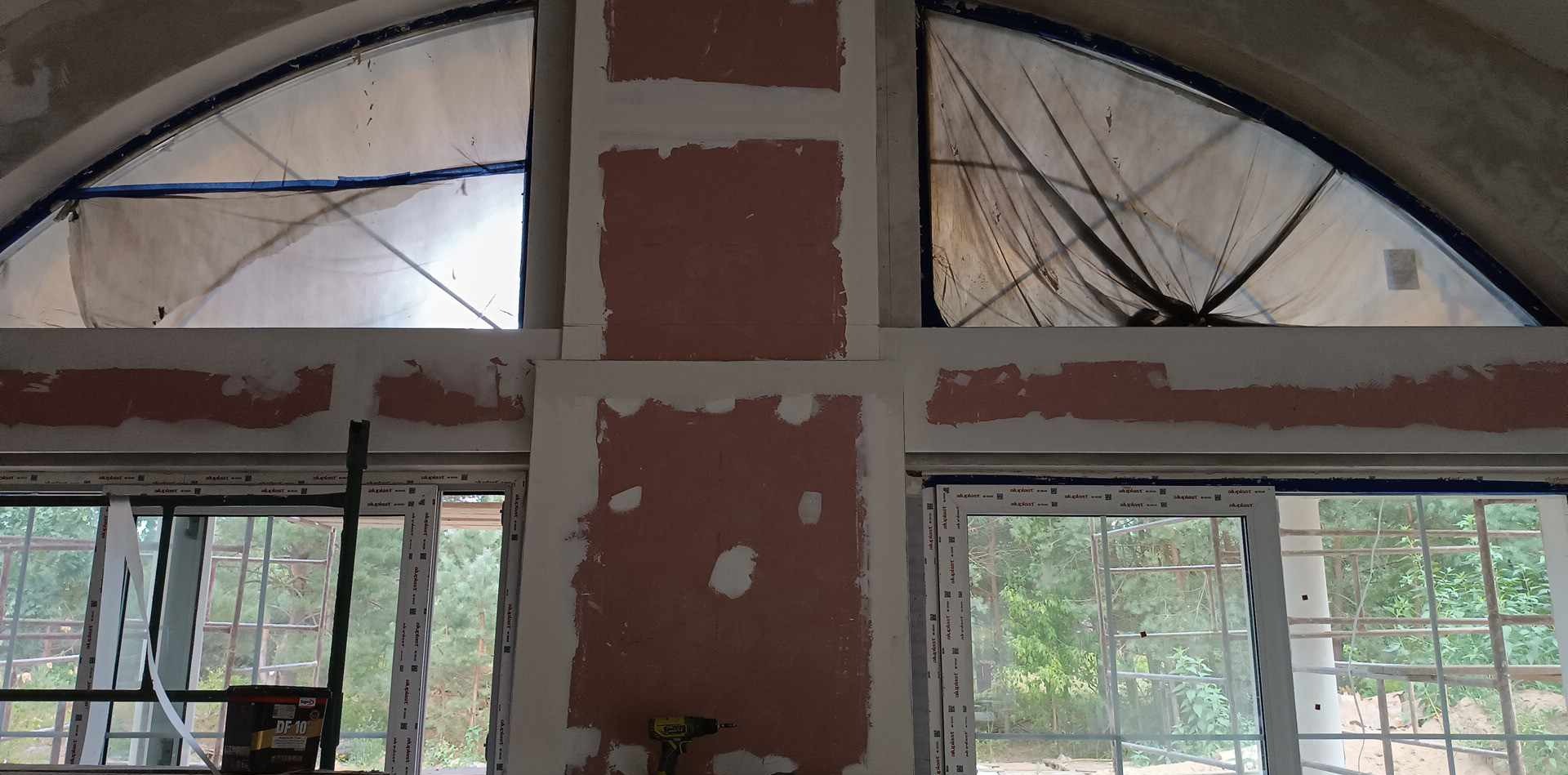 Wnętrze w trakcie prac wykończeniowych: ściany z częściowo nałożoną warstwą szpachli, okna zabezpieczone folią, widoczne narzędzia i wiadro z masą szpachlową.