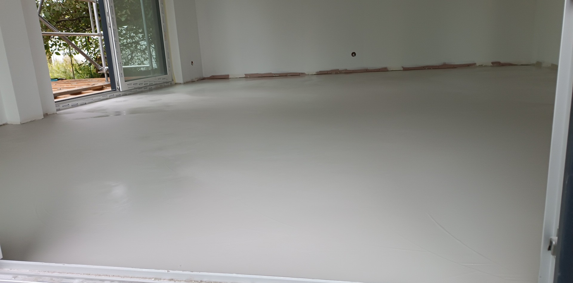 Wnętrze pokoju z wylewką samopoziomującą, widoczne ślady po wałku, przygotowane do dalszych prac wykończeniowych, okno balkonowe z widokiem na zewnątrz.