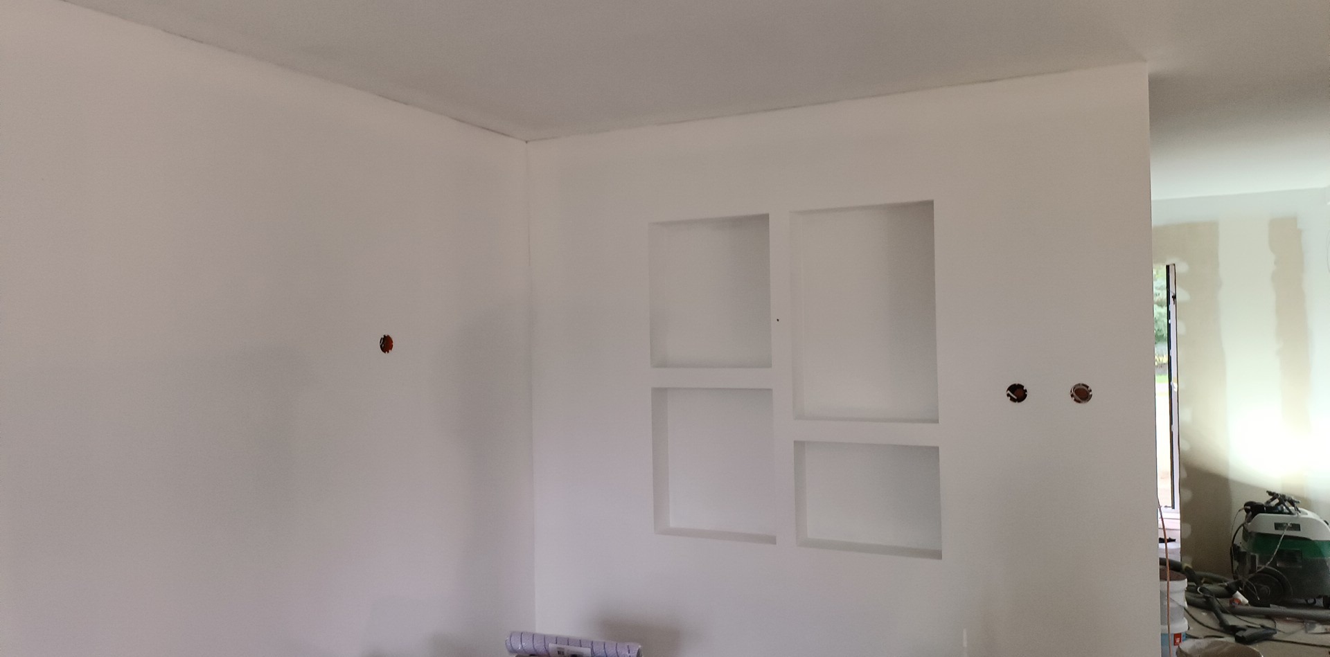 Świeżo pomalowane na biało wnętrze z wnęką na ścianie, widoczne puszki elektryczne i rolka tapety na podłodze.