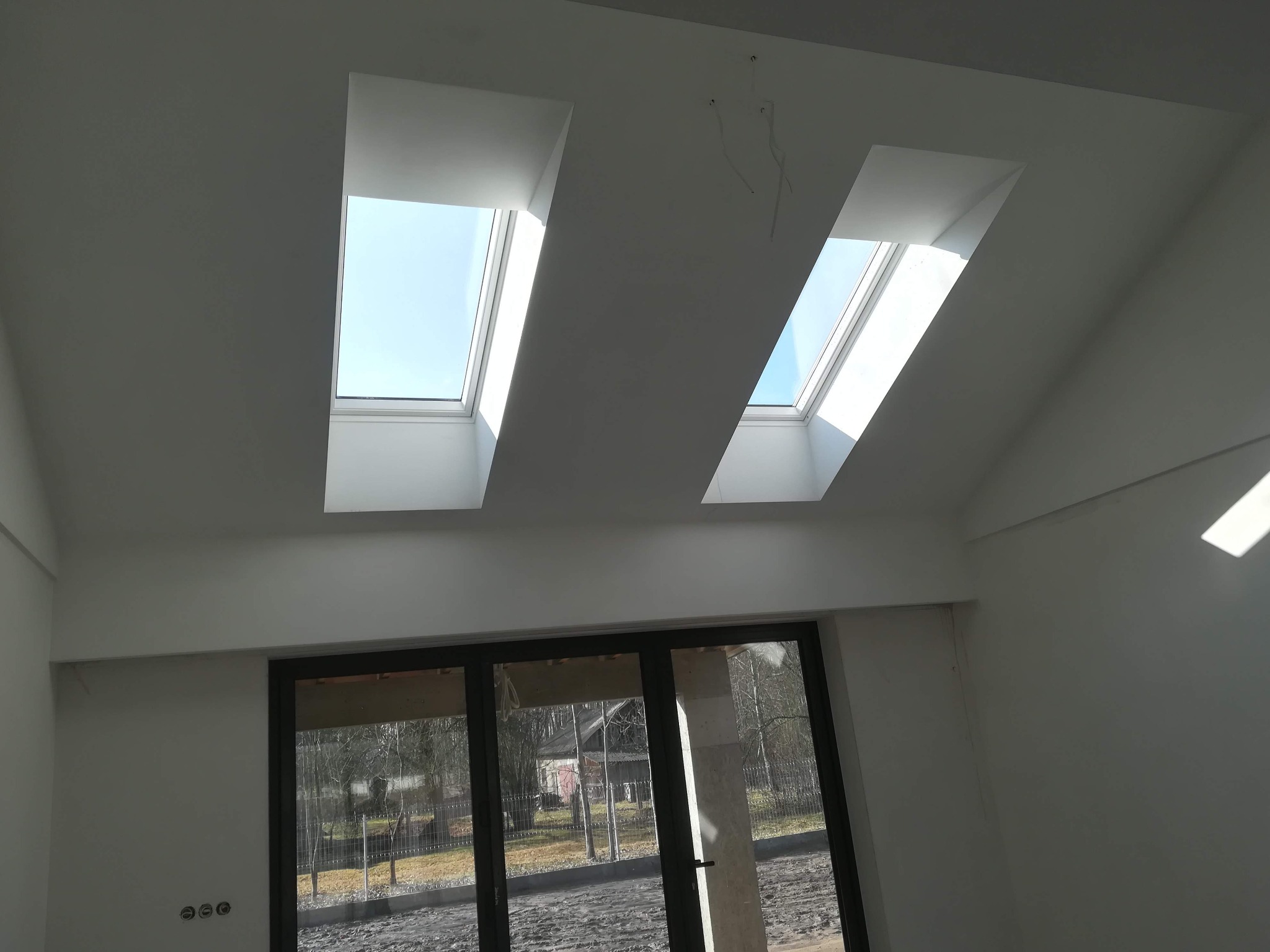 Wnętrze z oknami dachowymi i dużym oknem tarasowym, ściany przygotowane do dalszych prac wykończeniowych, widoczne wyprowadzone przewody elektryczne.