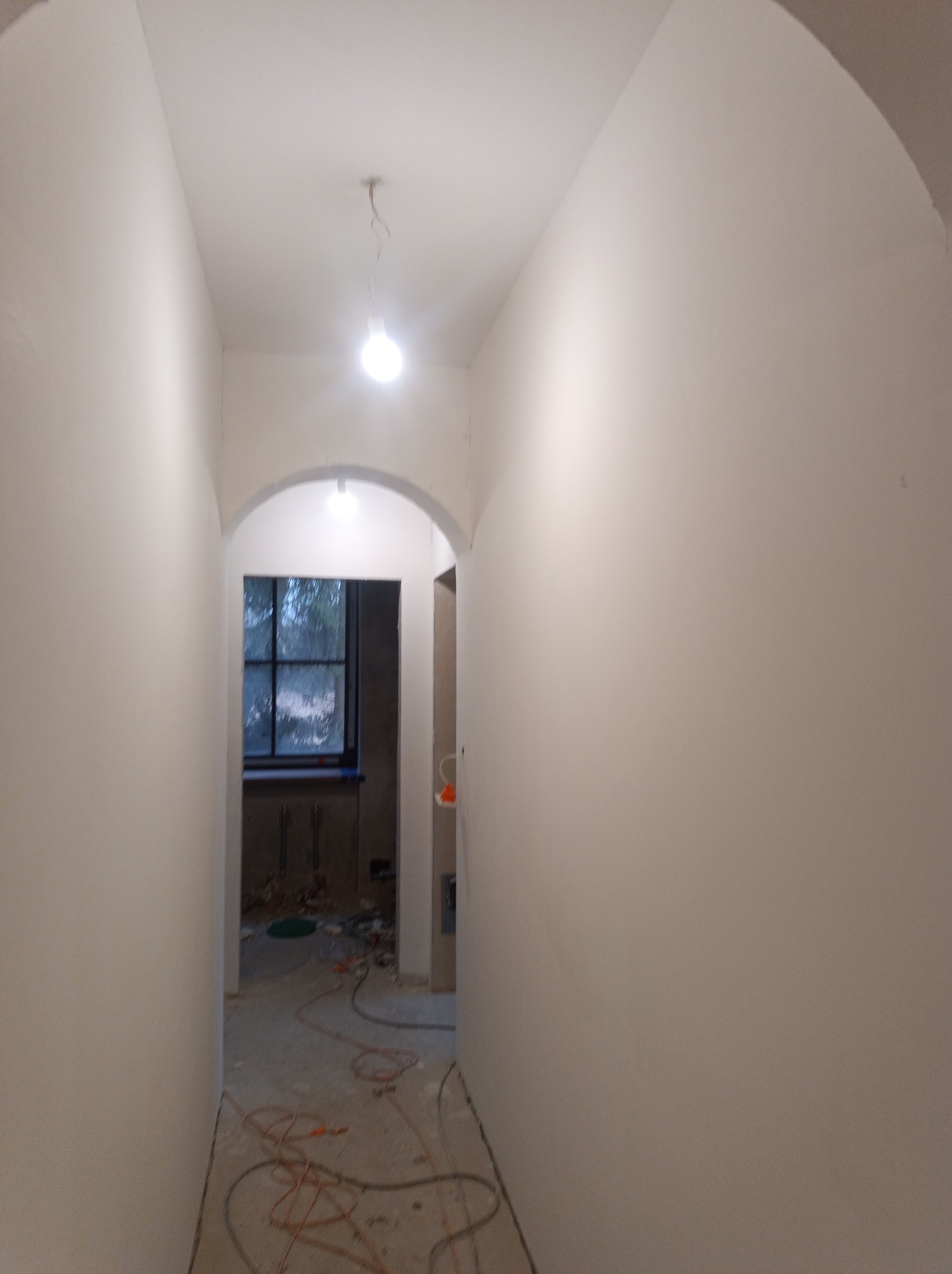 Korytarz w trakcie prac wykończeniowych, ściany pokryte gładzią, widoczne kable na podłodze i okno w głębi.