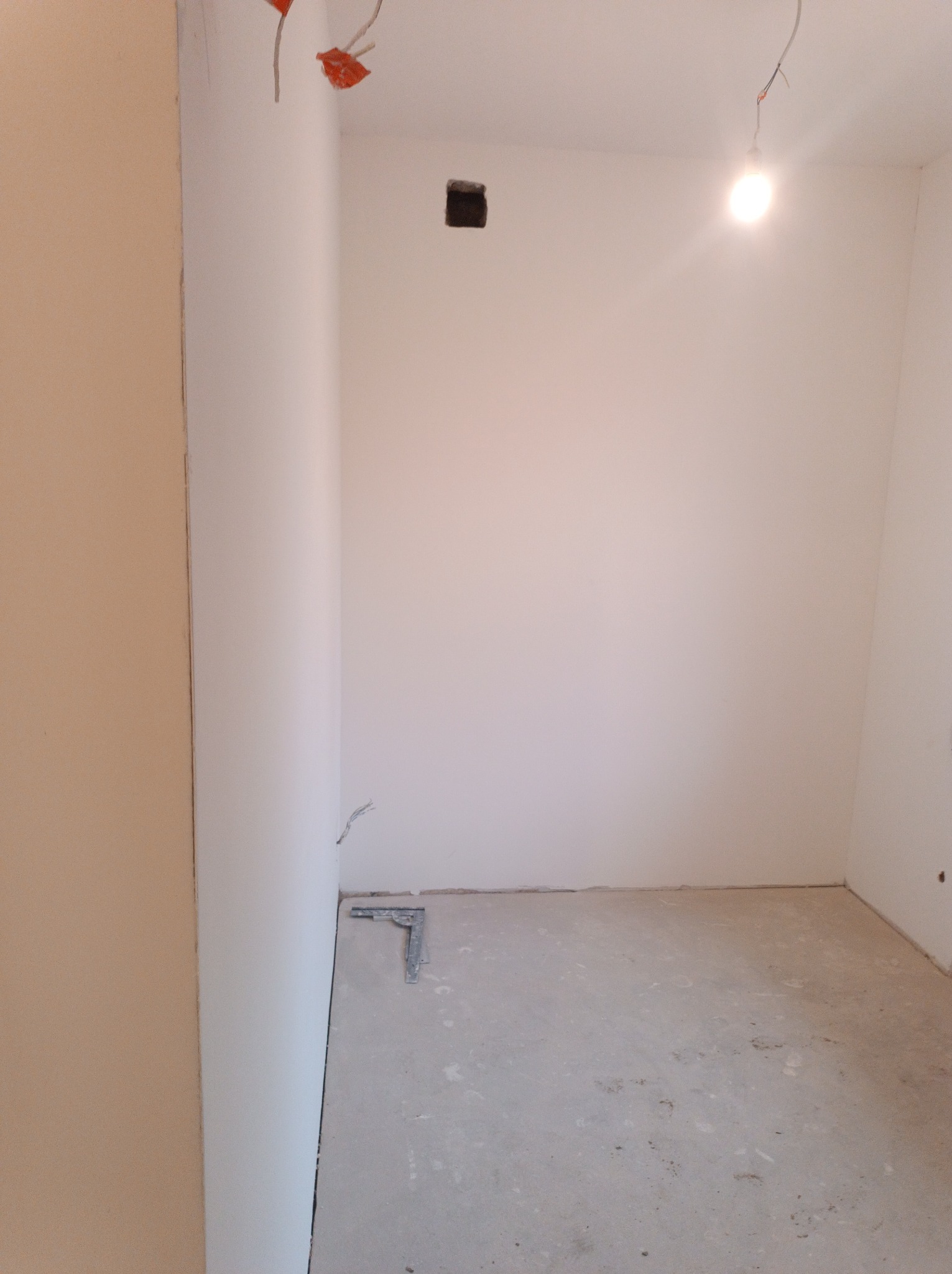 Pokój w trakcie prac wykończeniowych: białe ściany, surowy betonowy podkład, widoczne kable elektryczne, metalowy kątownik przy ścianie.