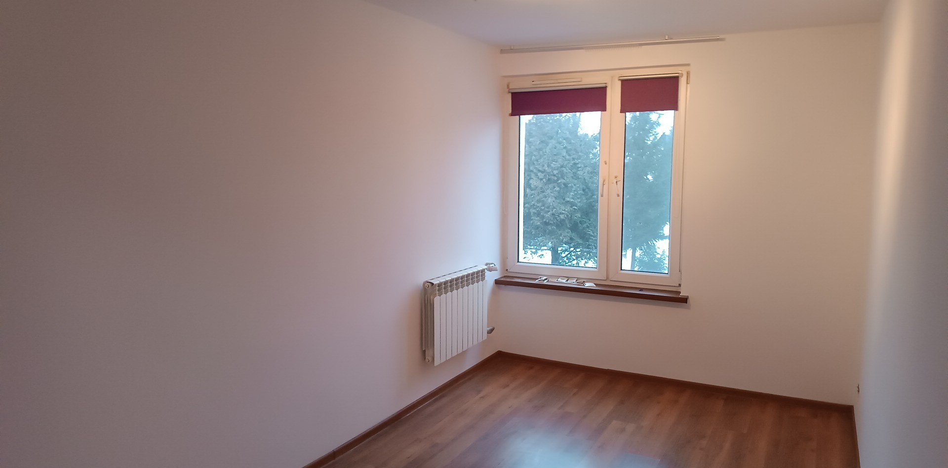 Pusty pokój z białymi ścianami, brązowymi panelami podłogowymi, grzejnikiem i oknem z bordowymi roletami.