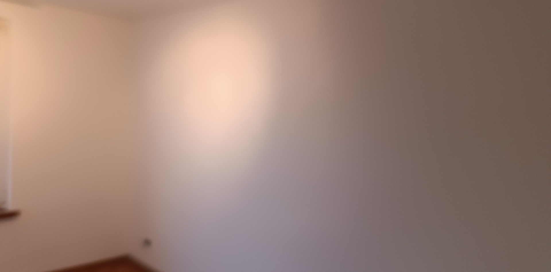 Nierównomiernie oświetlona, blado-różowa ściana w rogu pomieszczenia, widoczny fragment podłogi i okna.